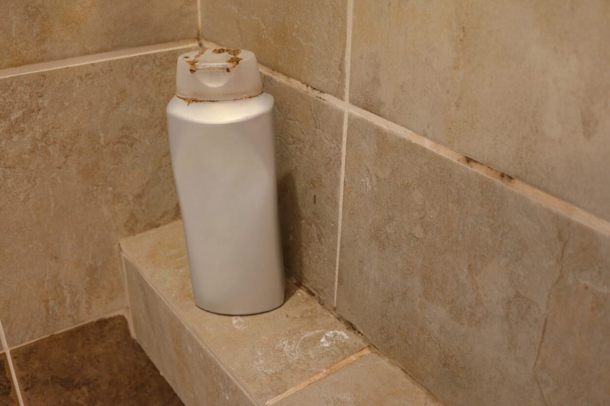 Shampoo bottle on a tiled shower shelf with orange residue in the grout and caulk